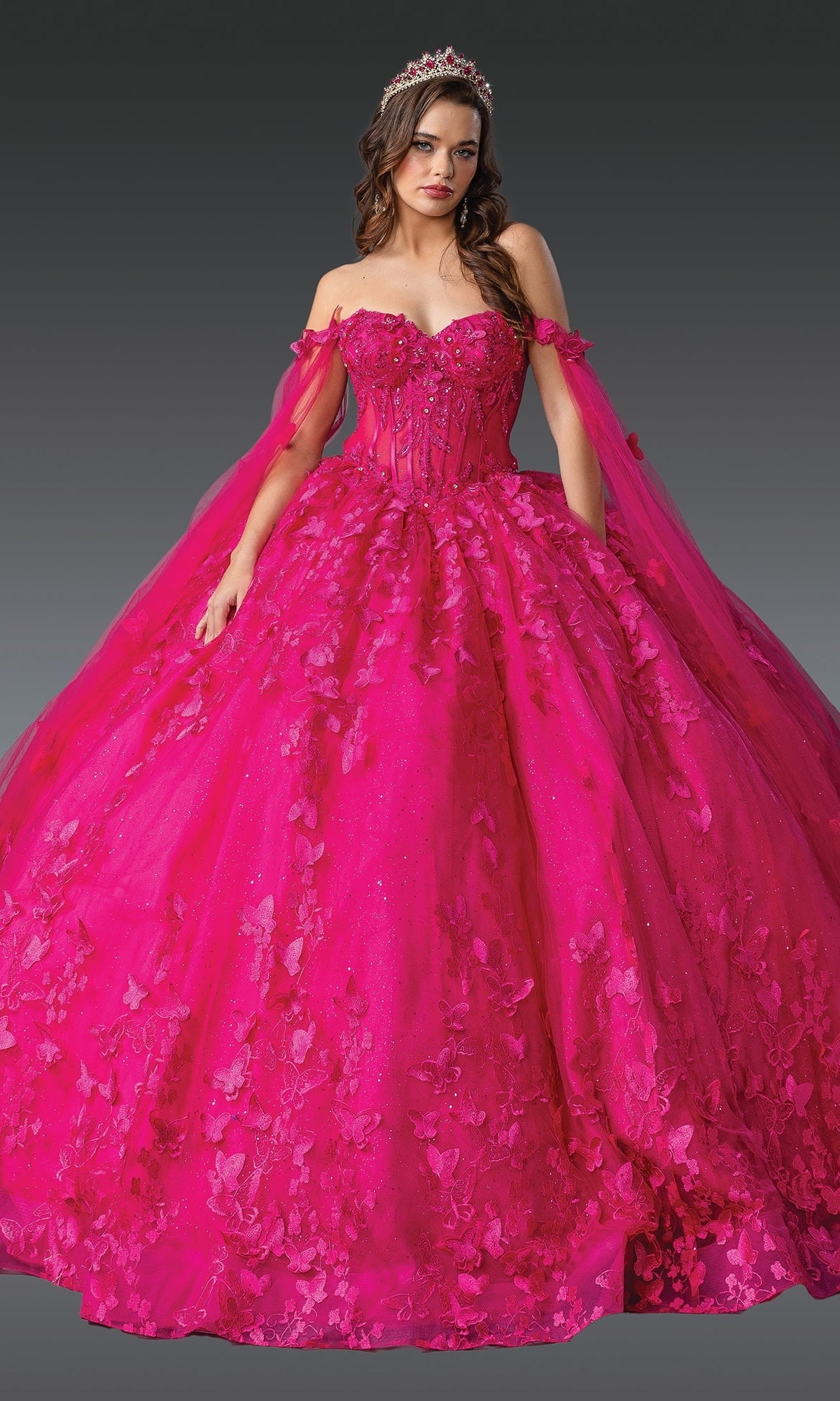A woman poses against a dark background in the Fuchsia Quinceanera Dress 1960 By Dancing Queen, featuring a corset bodice, floral appliqués, sheer cape, and tiara.